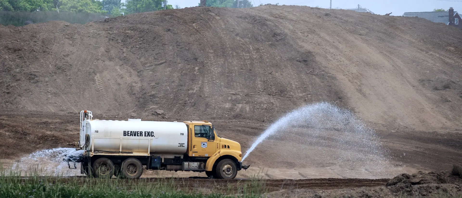 A water tanker truck spraying water across a bare earth embankment on a construction site